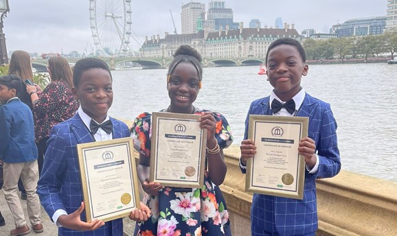 Two young boys and a girl in smart clothes are smiling holding awards with the london eye behind