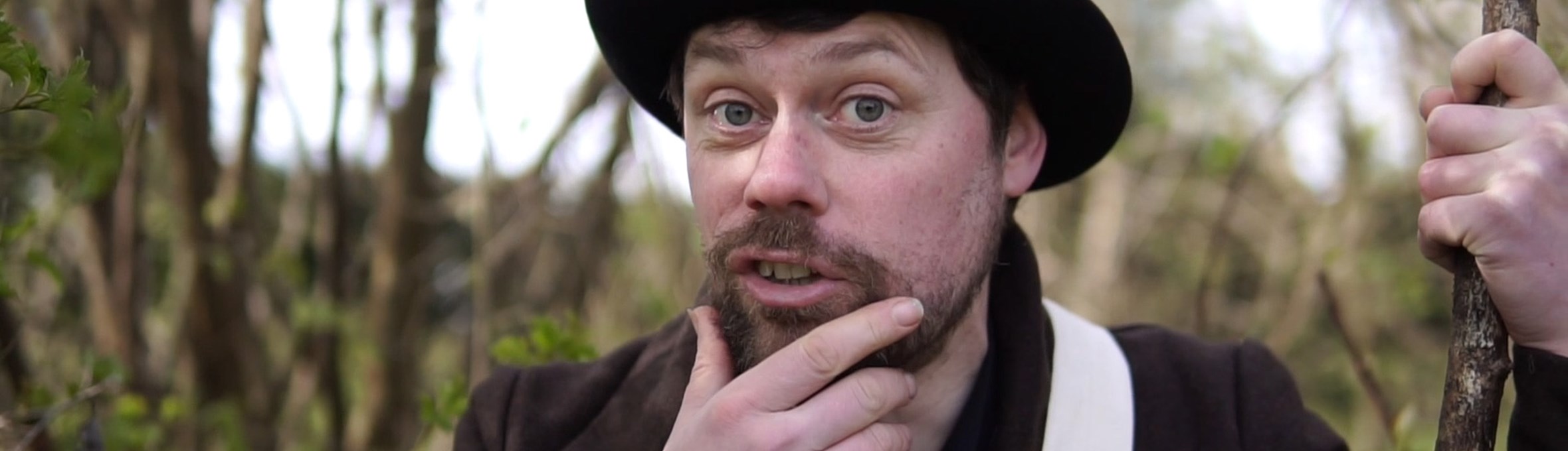 An actor is dressed in a small top hat and brown suit looking at the camera 