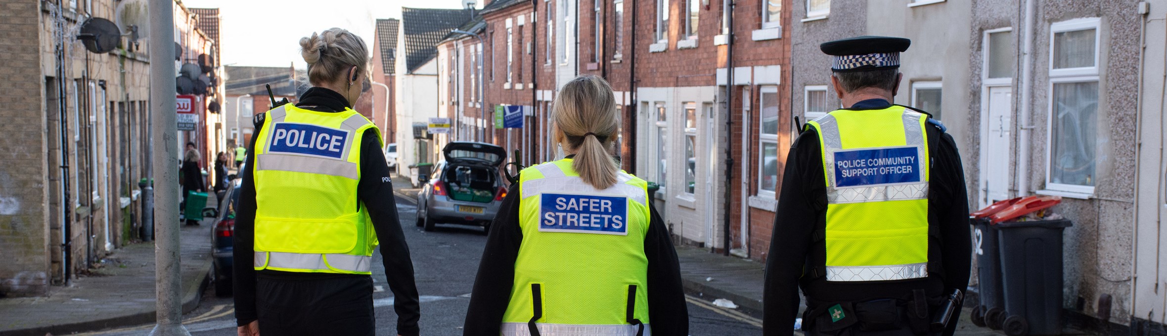 a police woman, a cpo and pcso wear high vis vests whilst walking through a street 